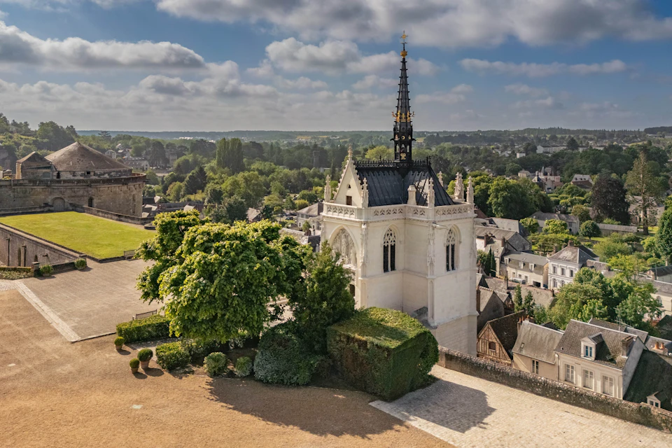 A historic chapel with a tall spire surrounded by greenery and overlooking a town under a partly cloudy sky.