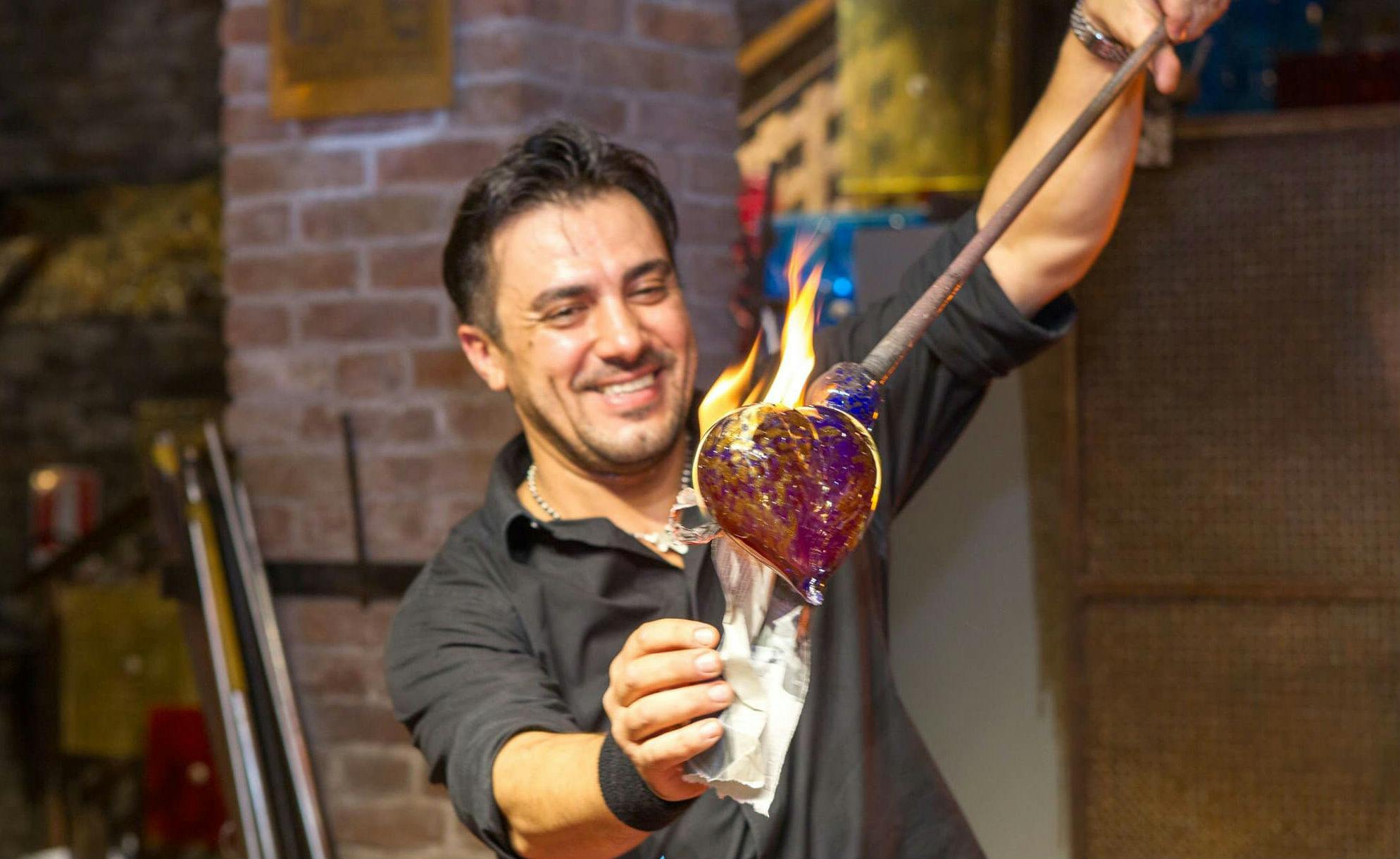 一位穿着黑色衬衫的微笑男子在玻璃吹制工坊里拿着一块发光的热玻璃，上面有火焰。