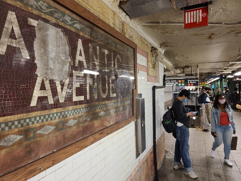 A subway station with a worn mosaic tile sign reading "Atlantic Avenue" and people waiting near the platform.