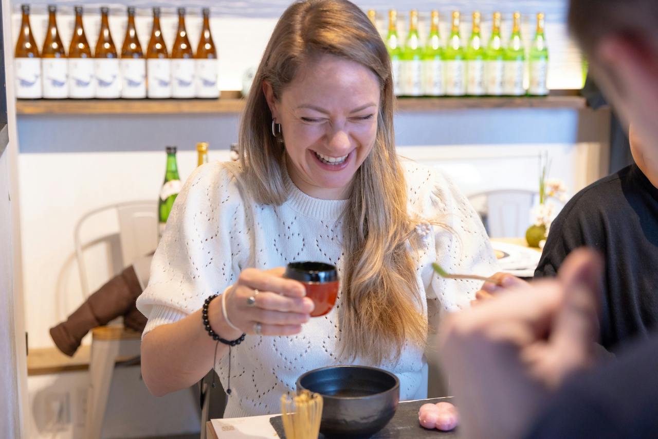 A woman is laughing while holding a small cup and sitting at a table with food items. Bottles are displayed in the background.