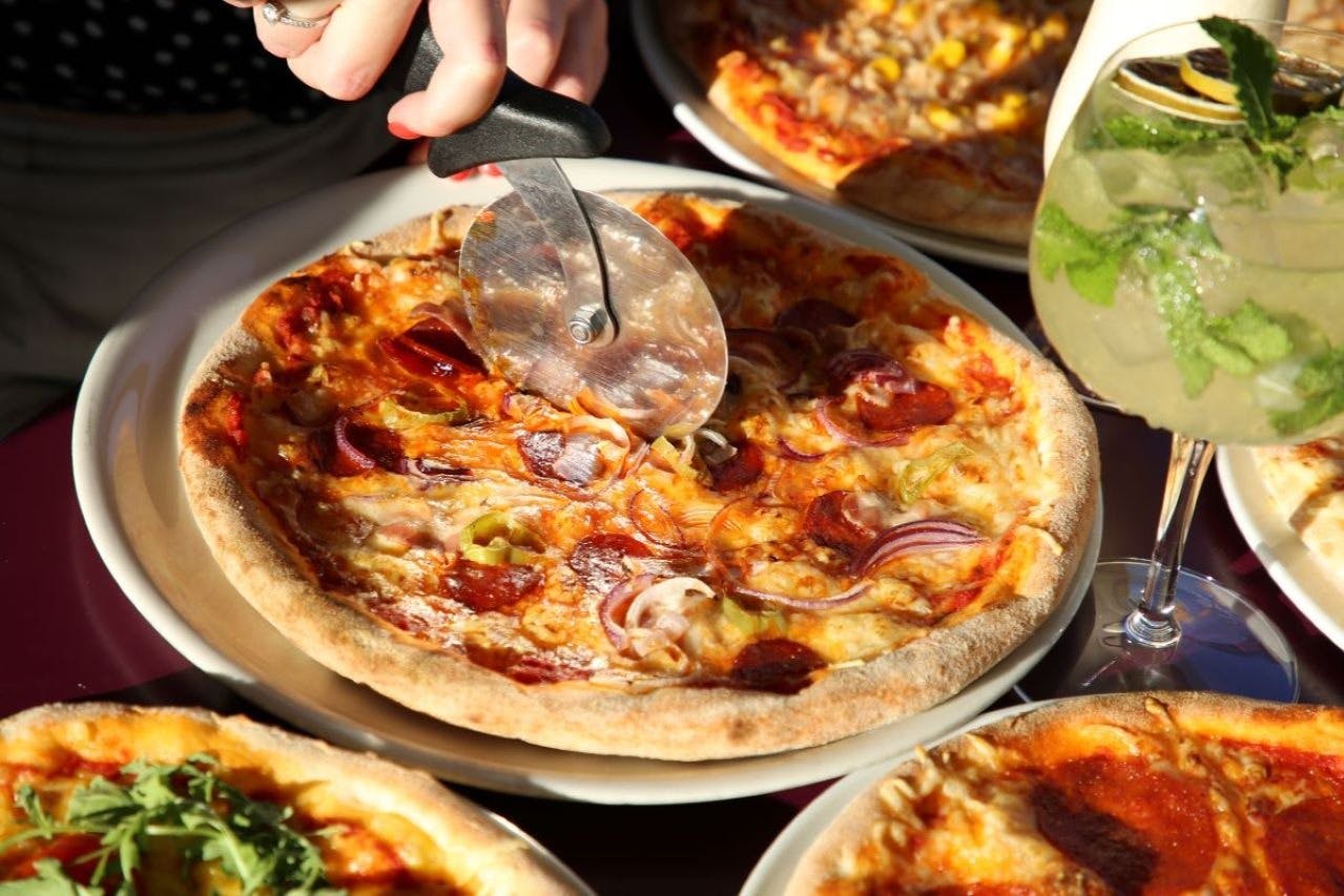 人はピザカッターを使って、ペパロニ、タマネギ、ピーマンをトッピングしたピザをスライスします。 他のピザやドリンクも近くにあります。