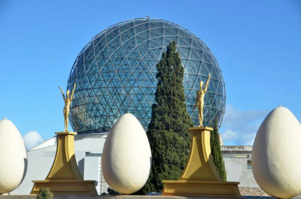 Geodesic dome behind statues holding eggs and a large egg sculpture, with tall trees and a blue sky in the background.