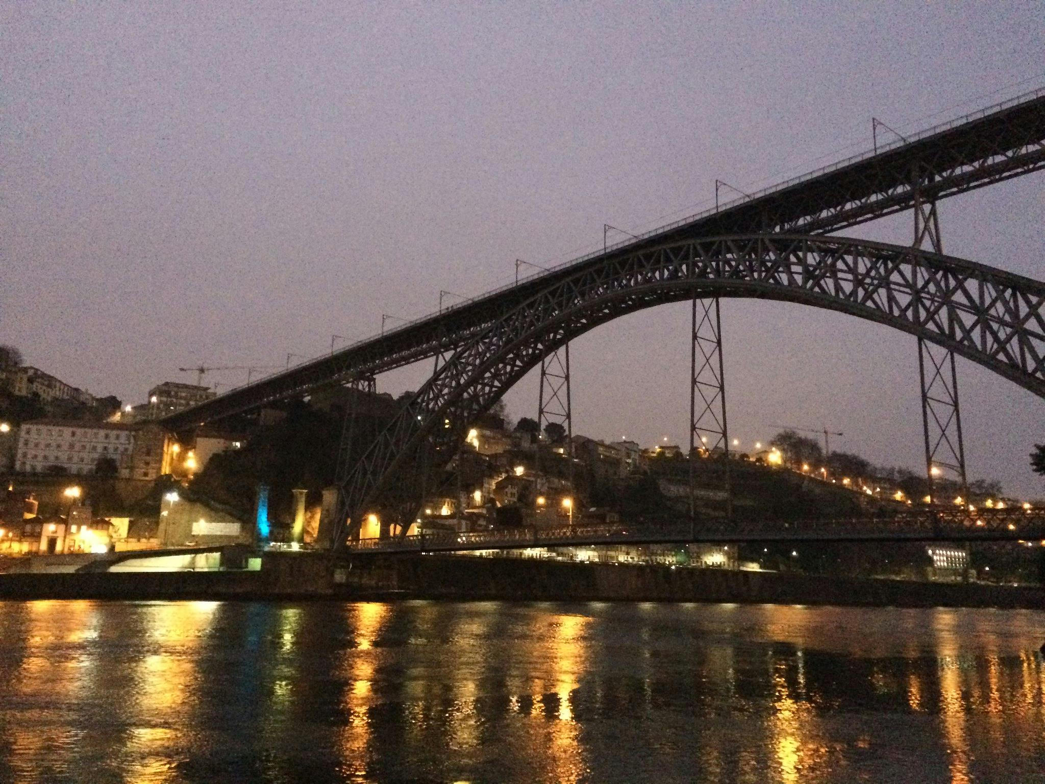 En upplyst bro sträcker sig över en flod i skymningen med stadsljus som reflekteras i vattnet och byggnader som är synliga på sluttningen.