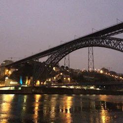 En upplyst bro sträcker sig över en flod i skymningen med stadsljus som reflekteras i vattnet och byggnader som är synliga på sluttningen.