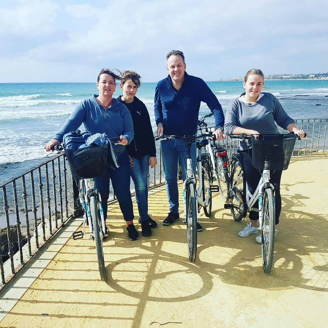 Four people stand beside their bicycles on a promenade by the sea, with waves and a coastal town in the background.