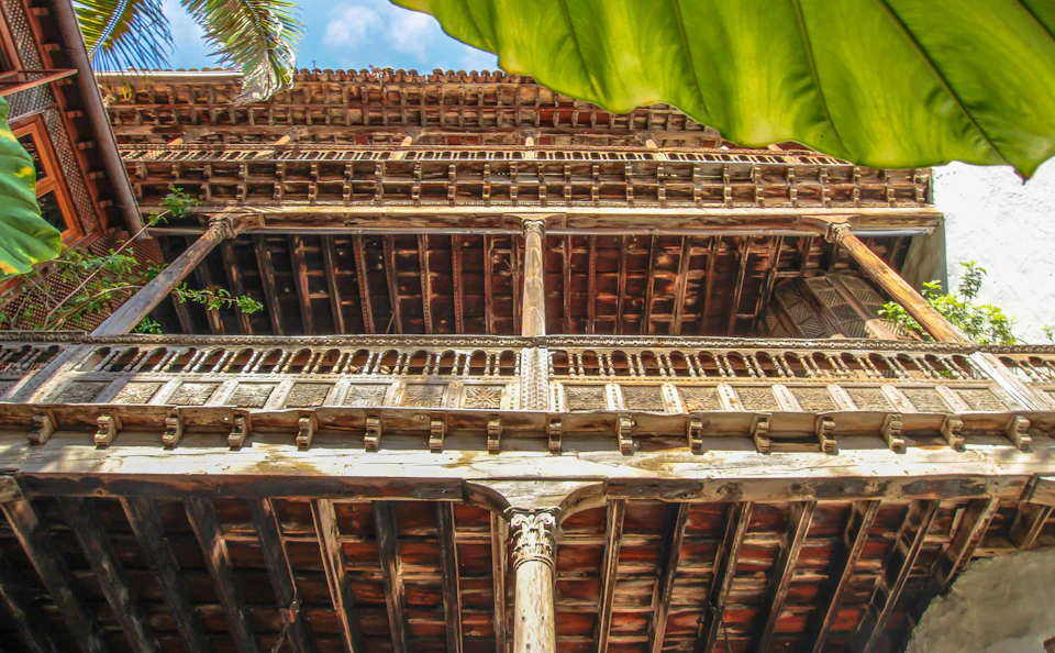 View from below of an intricate, multi-story wooden building with balconies, framed by a large green leaf and a palm frond.