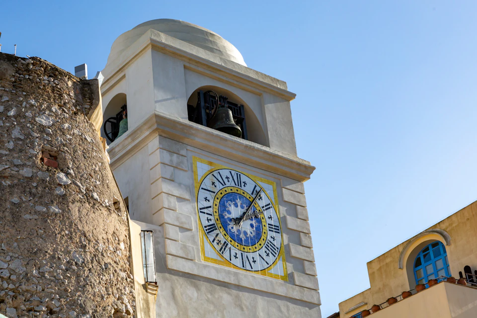A tall clock tower with a decorative clock face and a bell inside its top structure, against a clear blue sky.