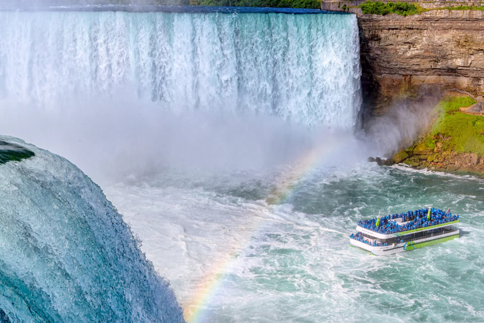 A boat full of people in blue raincoats approaches a large waterfall, with a rainbow visible in the mist.