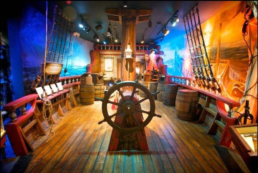 Interior of a brightly lit pirate-themed exhibit with a large ship's wheel, wooden barrels, ropes, and colorful murals.