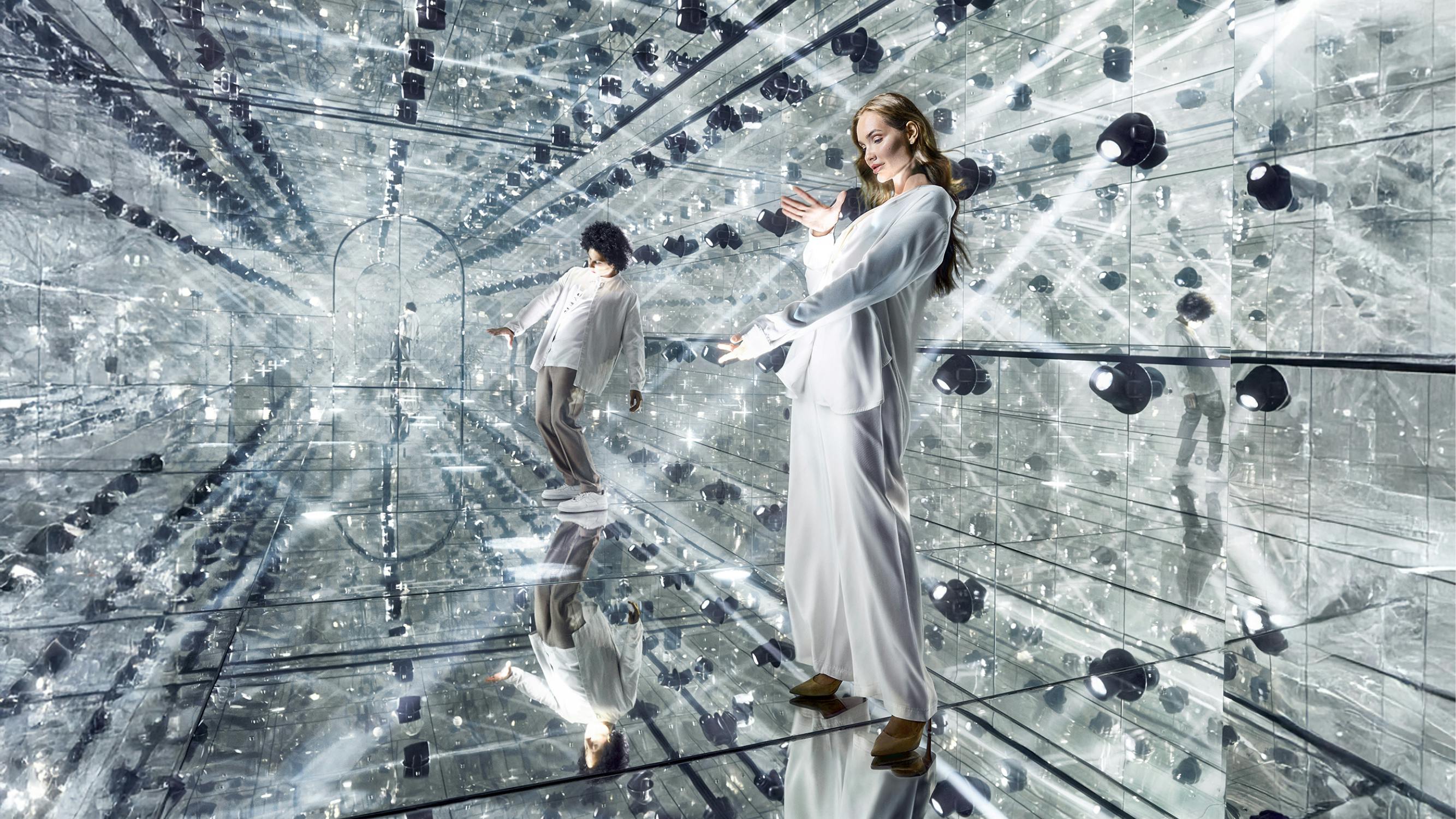 Δύο άτομα με λευκά ρούχα ποζάρουν σε ένα δωμάτιο με καθρέφτες στους τοίχους και τα πατώματα, δημιουργώντας ένα καλειδοσκοπικό αποτέλεσμα με πολλαπλές αντανακλάσεις.