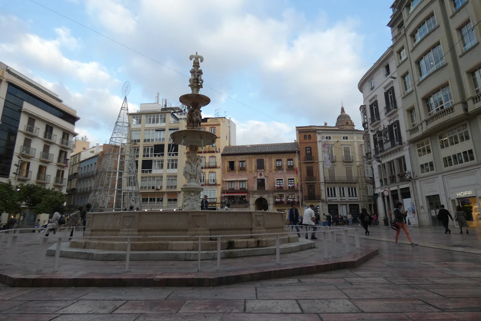 A plaza with a central fountain, surrounded by historic and modern buildings, people walking, and a metal framework structure.