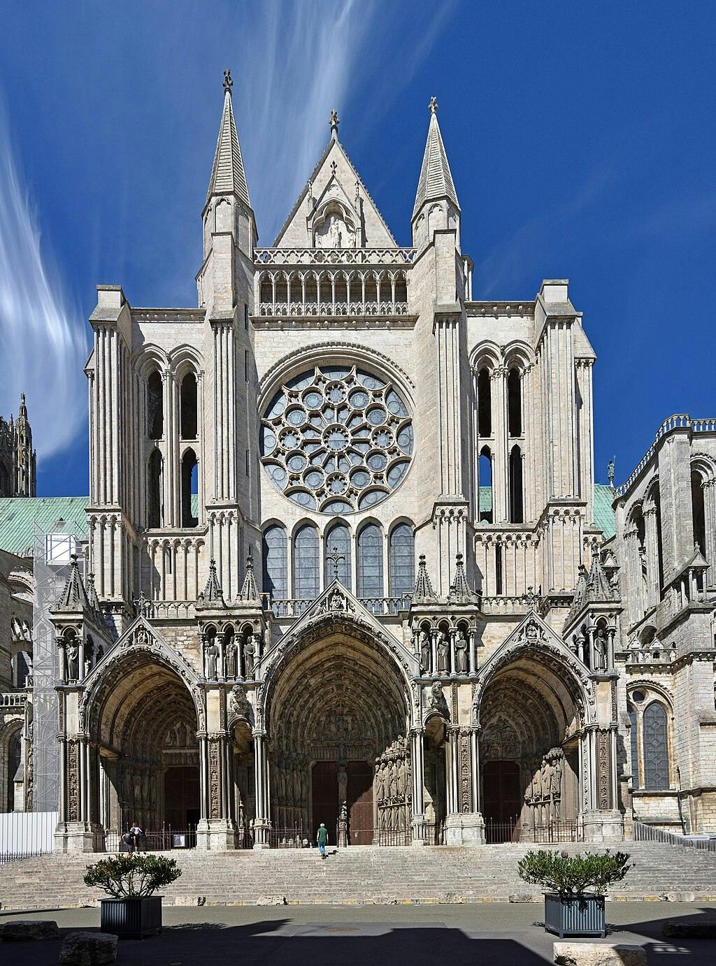 Gothic cathedral facade with detailed arches, rose window, and twin spires under a clear blue sky; a person stands on the steps.