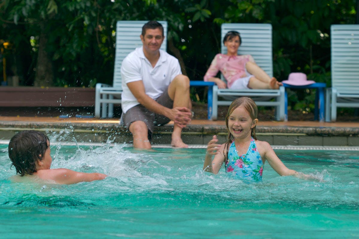 Two children play in a pool while two adults relax on lounge chairs in the background. Trees and greenery are visible behind them.