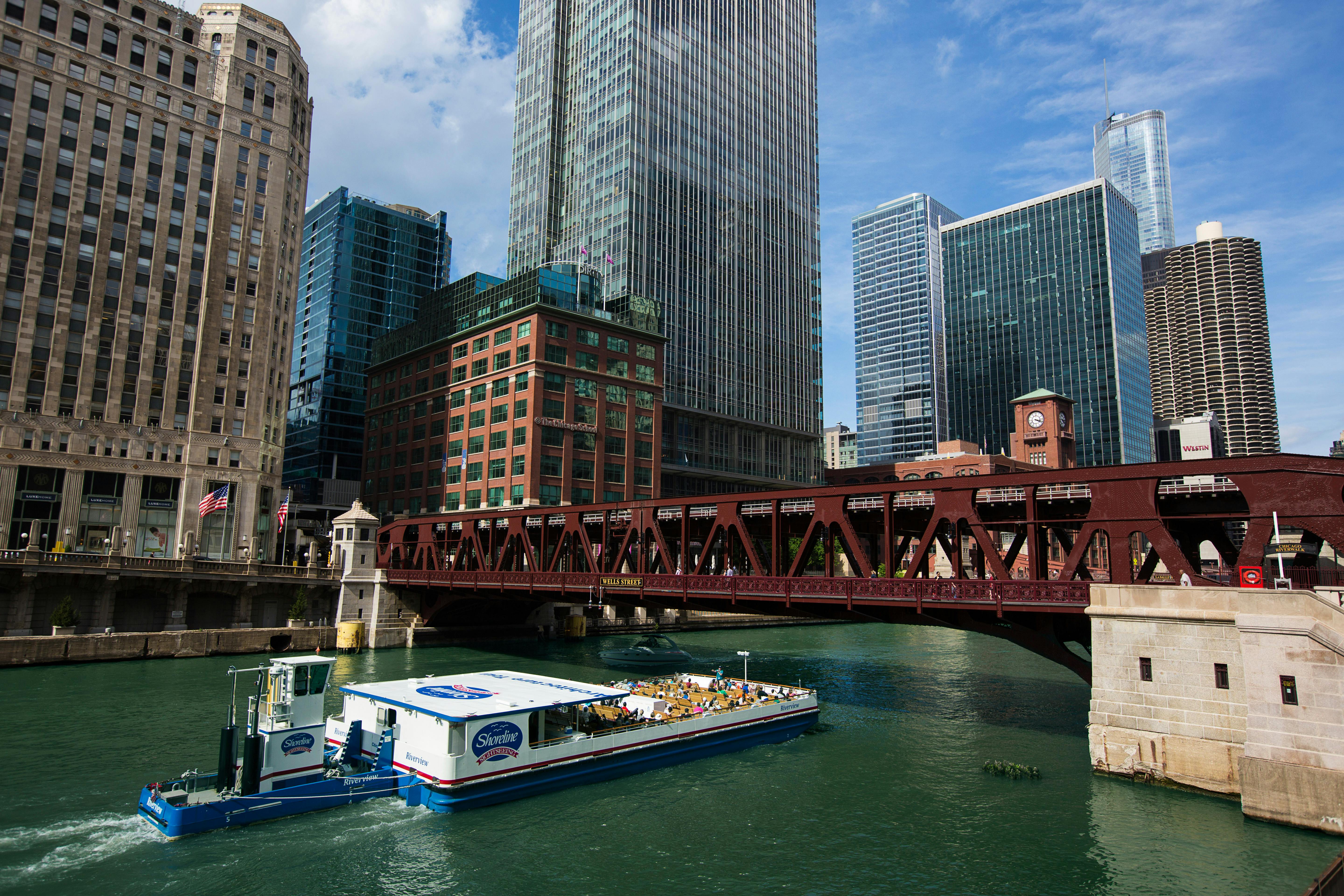 Una barca in tour in barca su un fiume sotto un ponte con alti edifici moderni sullo sfondo.