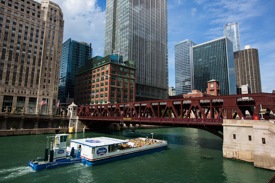 A boat cruising on a river under a bridge with tall, modern buildings in the background.