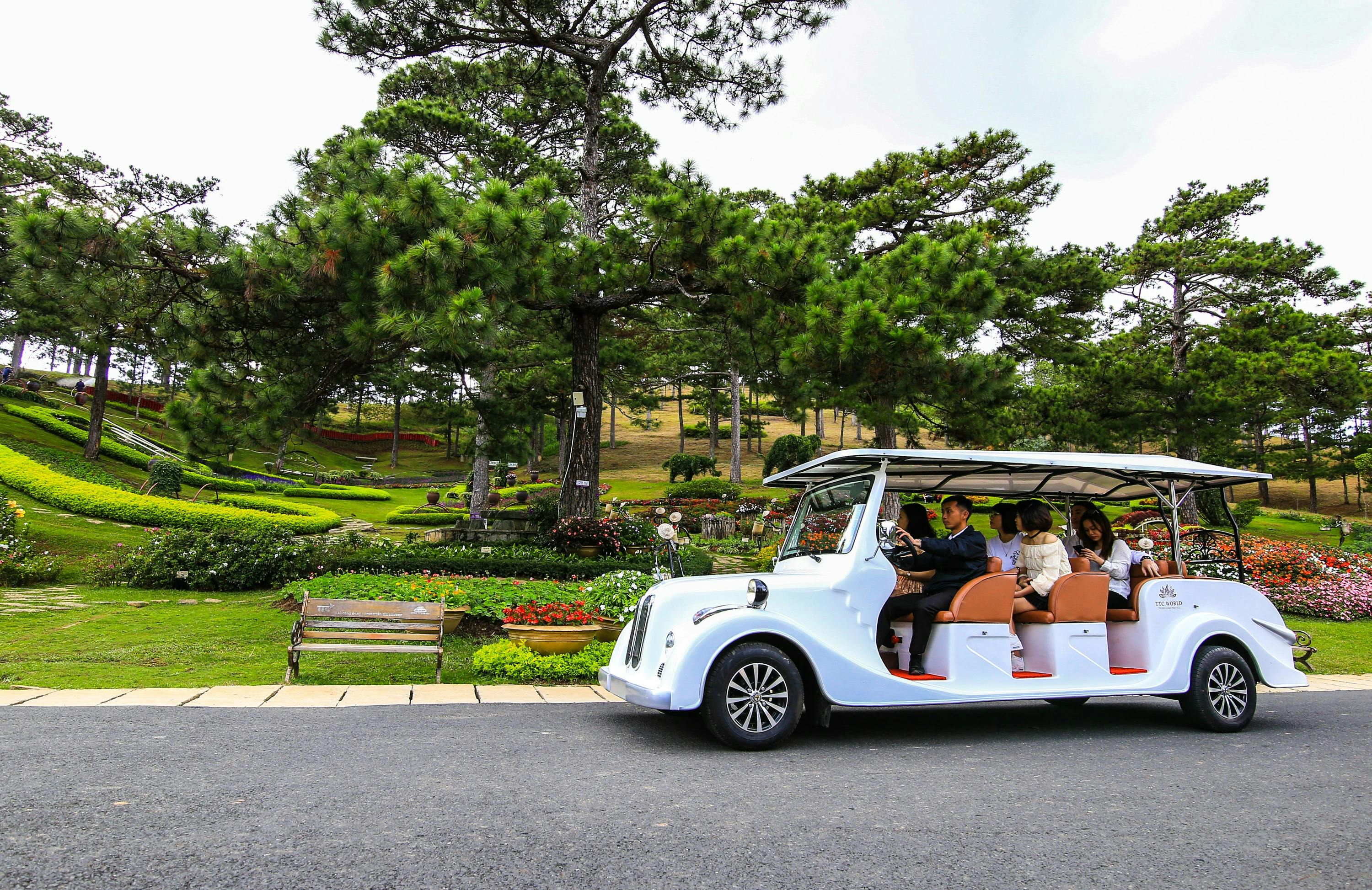 A white open-air vehicle with people inside driving past a landscaped garden with trees and flowers on a sunny day.