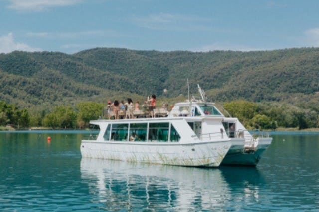 Un vaixell blanc amb gent a la coberta superior navega per un llac tranquil amb verdes turons boscosos al fons.