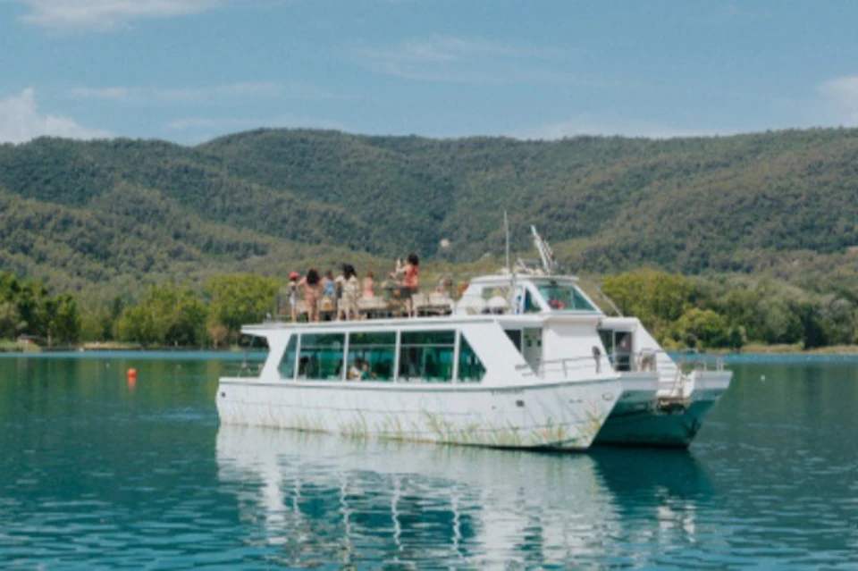 A white boat with people on the upper deck is sailing on a calm lake with green wooded hills in the background.
