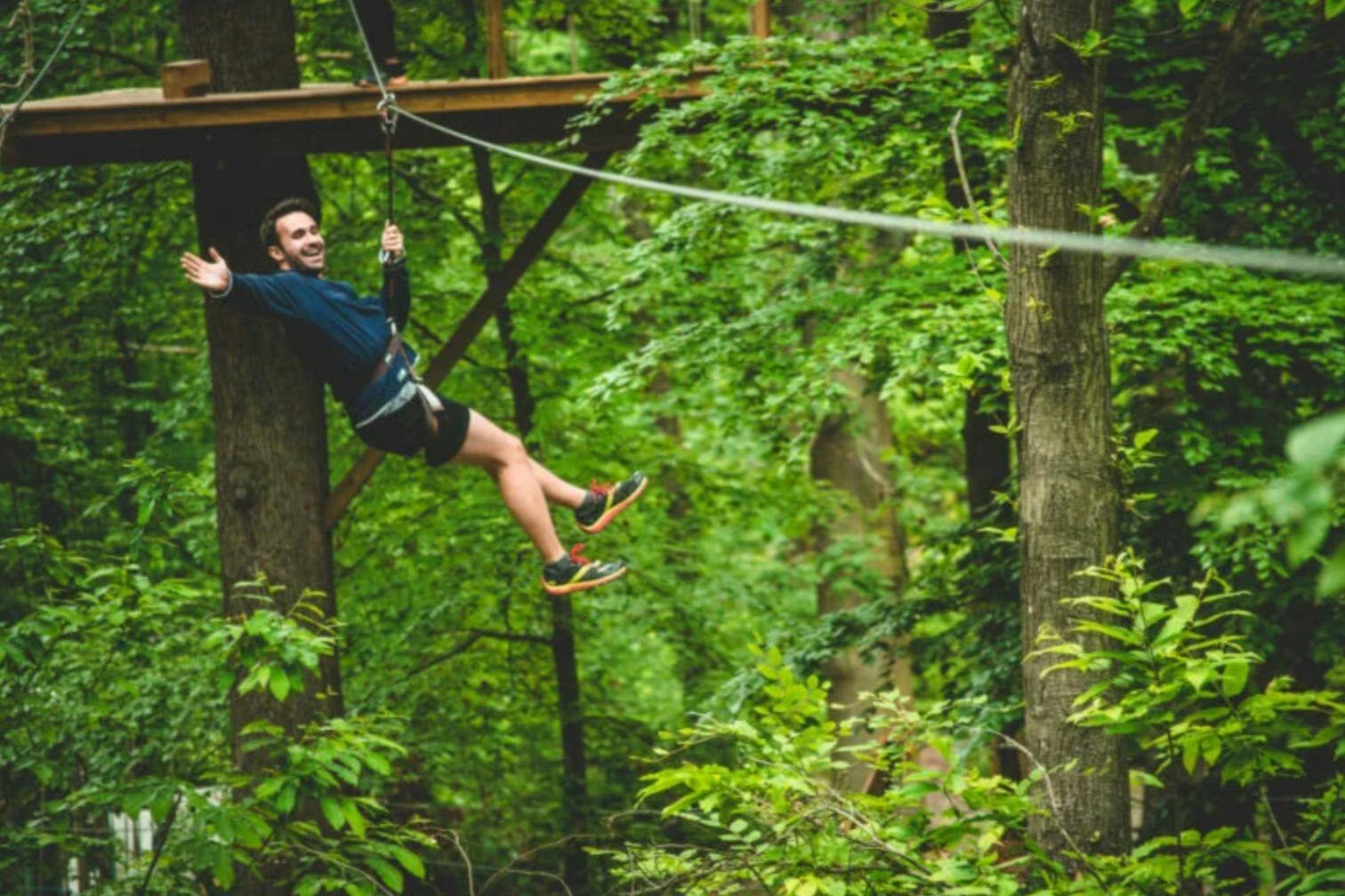 Una persona somriu mentre es desplaça en tirolina per un bosc exuberant, amb una plataforma de fusta i un folre verd de fons.