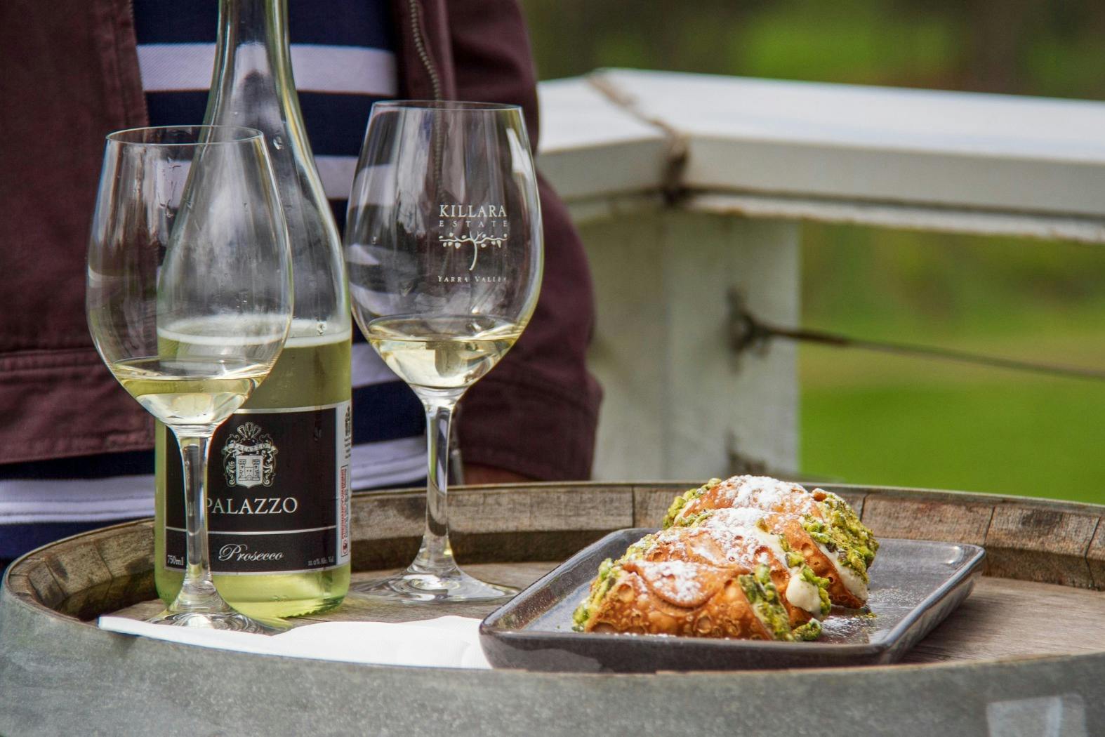 A table with a bottle of wine, two glasses, and a plate of pastries dusted with powdered sugar outdoors.