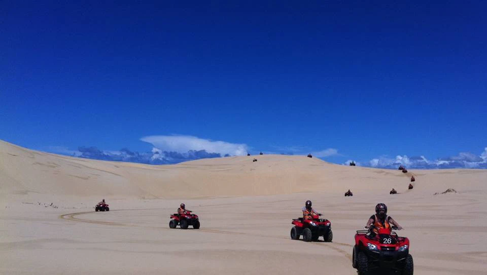 People riding red ATVs across a vast, sandy desert under a bright blue sky.