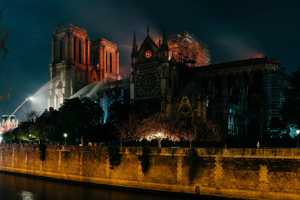 A nighttime view of the illuminated Notre Dame Cathedral with scaffolding, trees, and a riverbank in the foreground.