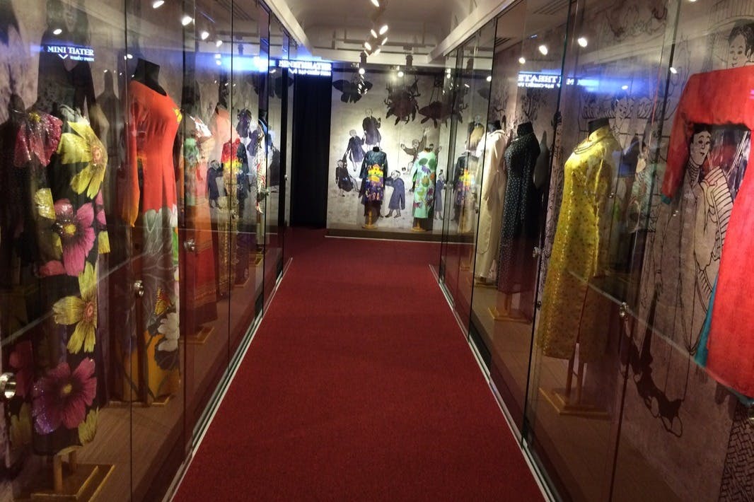 A hallway with red carpet, flanked by glass displays showcasing mannequins dressed in colorful vintage clothing.