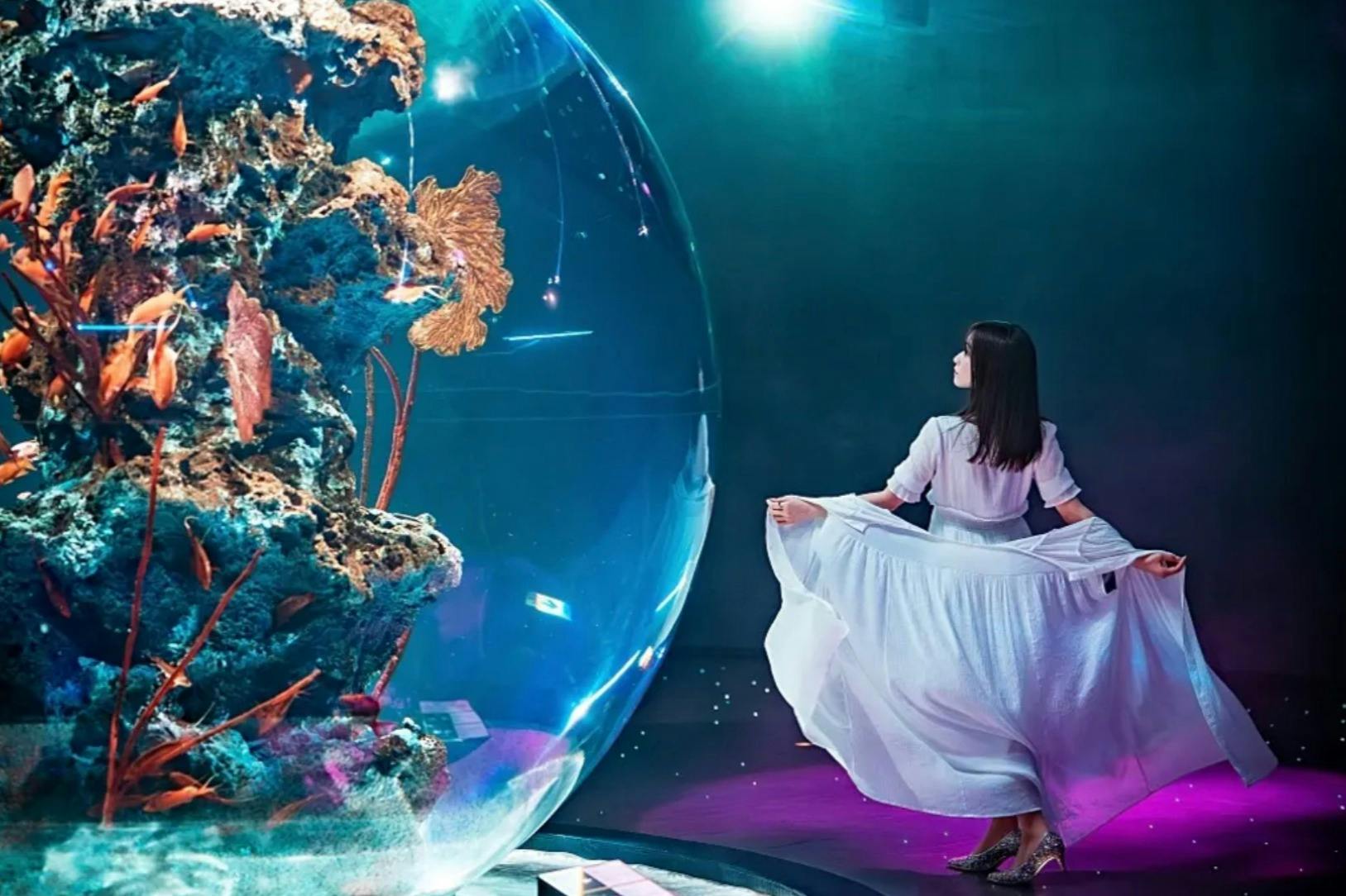 Kobieta w płynącej białej sukience stoi w pobliżu dużego akwarium z kolorowym życiem morskim, pod dramatycznym oświetleniem.