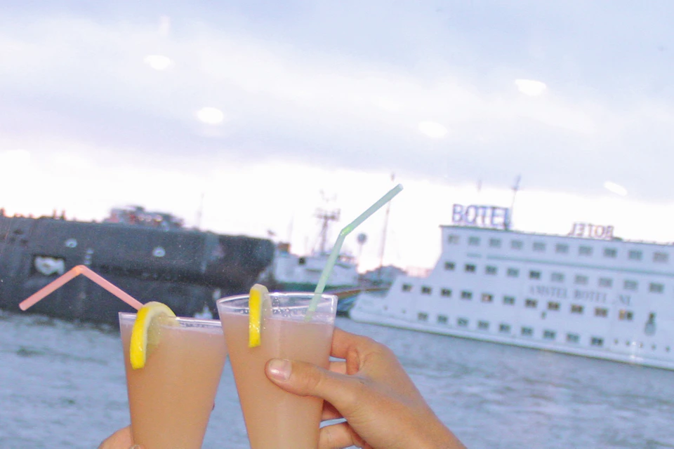 Woman smiling, dining at a riverside restaurant, holding a cocktail. Plates of pancakes and condiments on the table. River and boats outside.