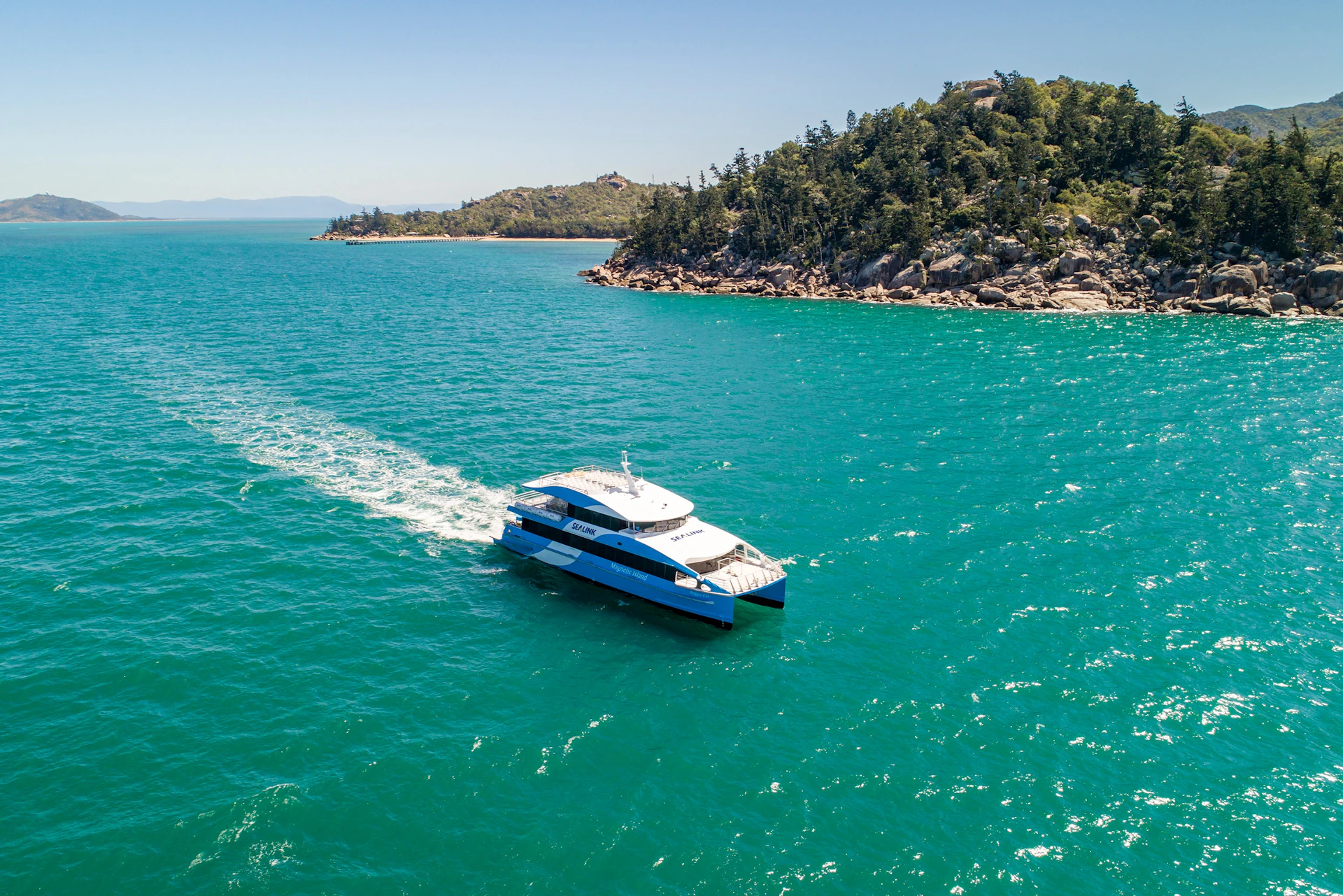 A blue and white catamaran sailing on clear turquoise water near a forested shoreline under a clear sky.