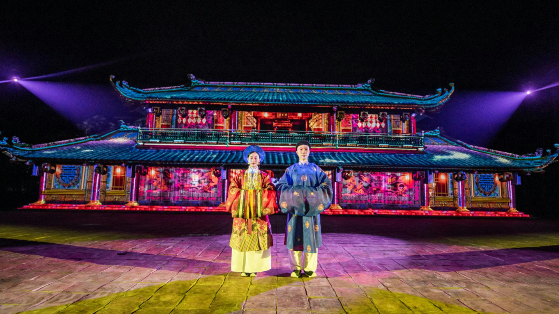 Two people in traditional clothing stand in front of an illuminated, colorful building at night.