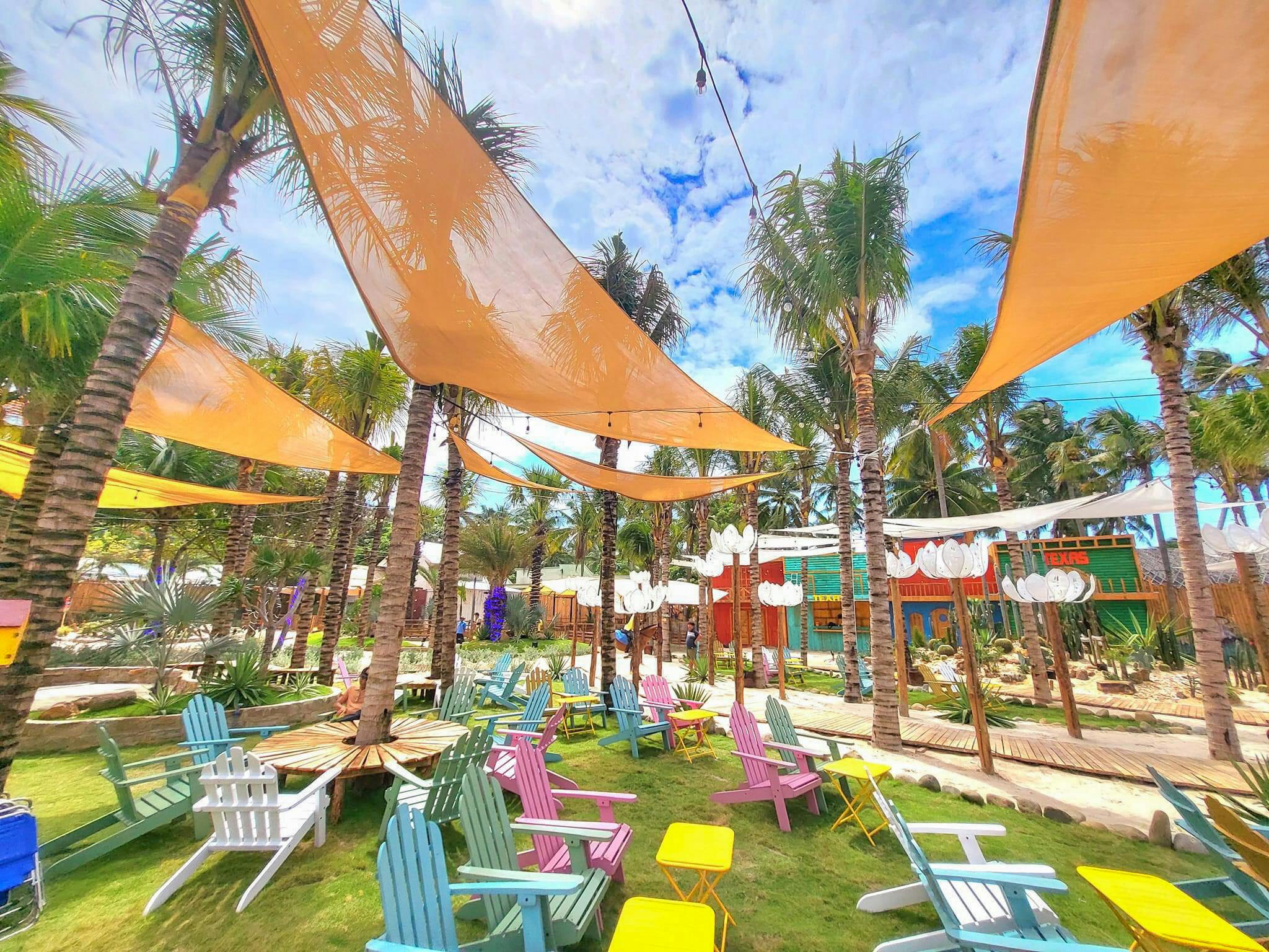 A tropical outdoor seating area with colorful chairs, yellow tables, palm trees, and orange fabric canopies under a sunny sky.