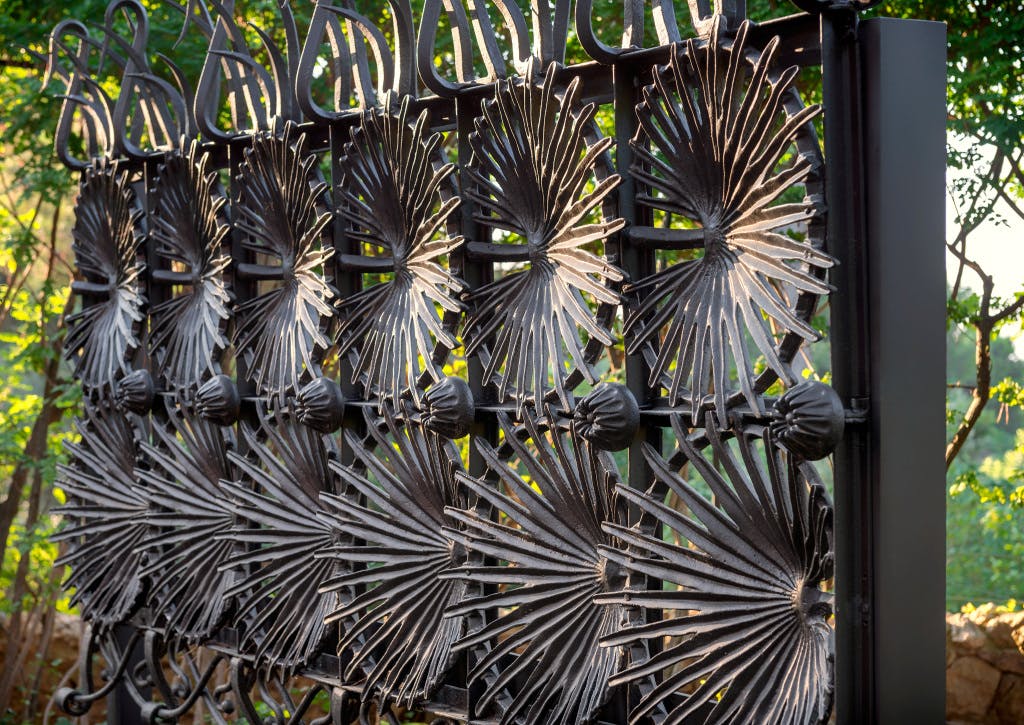 ファンのようなパターンを特徴とする複雑に設計された錬鉄製のゲート。隙間を通って太陽光がフィルタリングされ、背景には緑があります。