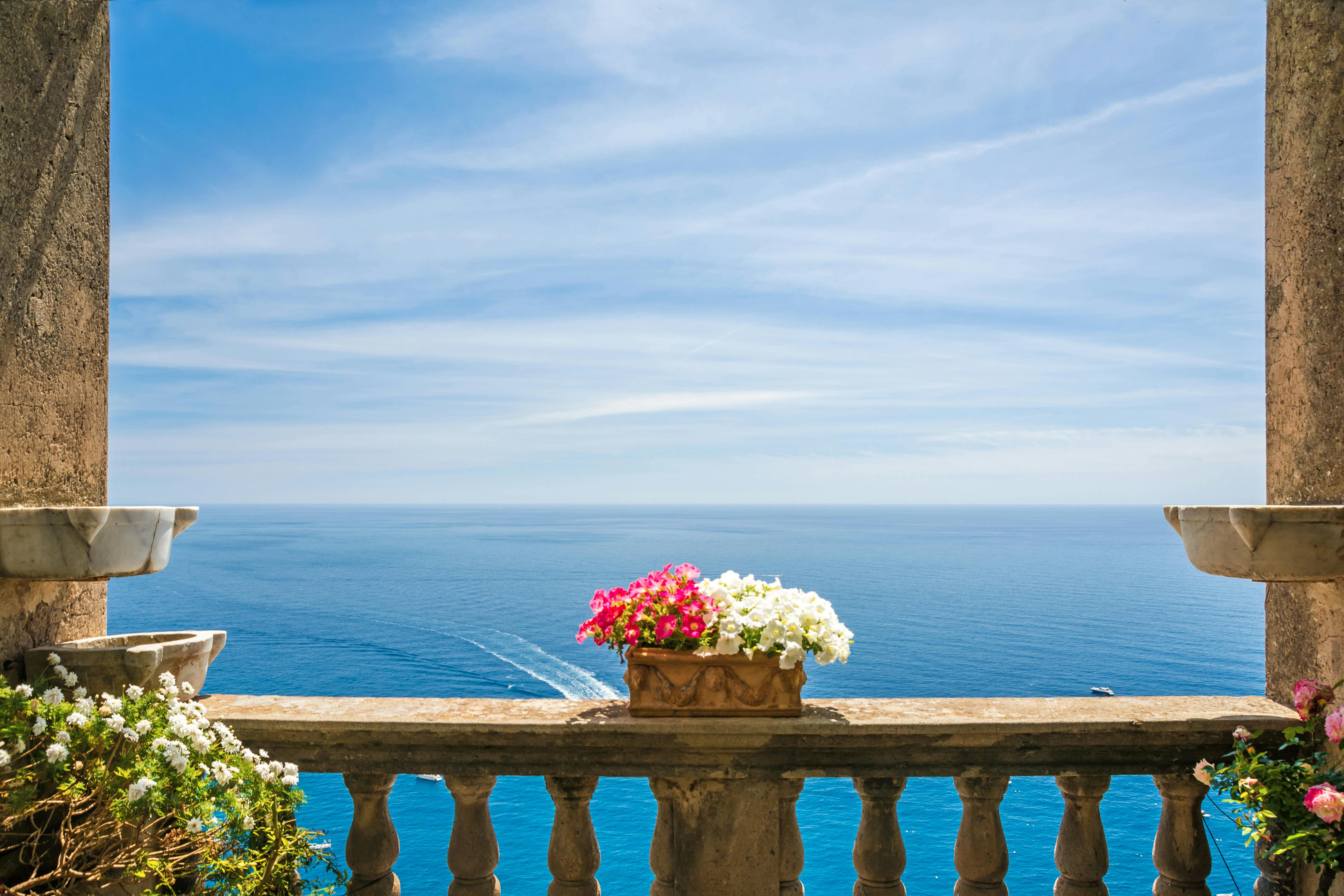 Stone balustrade with a flower pot containing pink and white flowers, overlooking a calm blue sea under a bright, clear sky.
