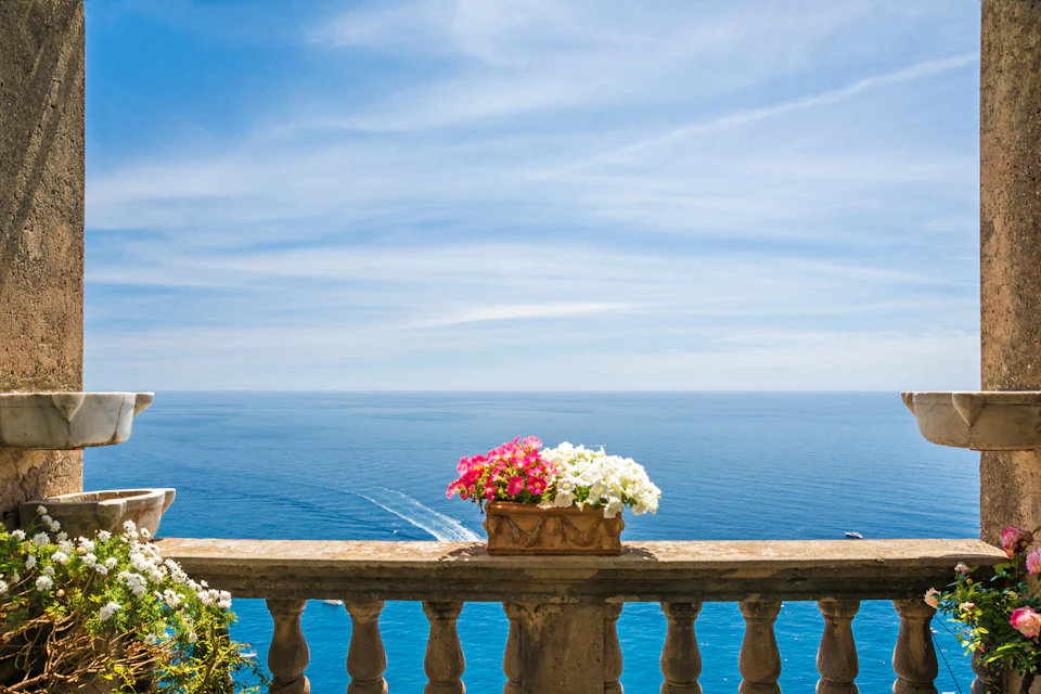 Stone balustrade with a flower pot containing pink and white flowers, overlooking a calm blue sea under a bright, clear sky.