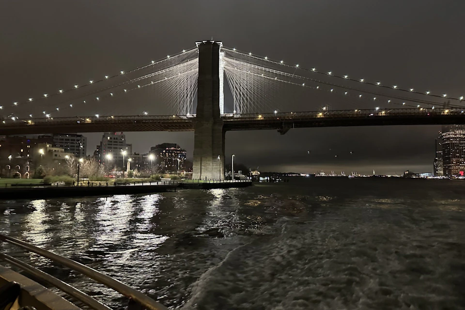 A nighttime view of a lit suspension bridge over a calm river, with reflections on the water and buildings in the background.