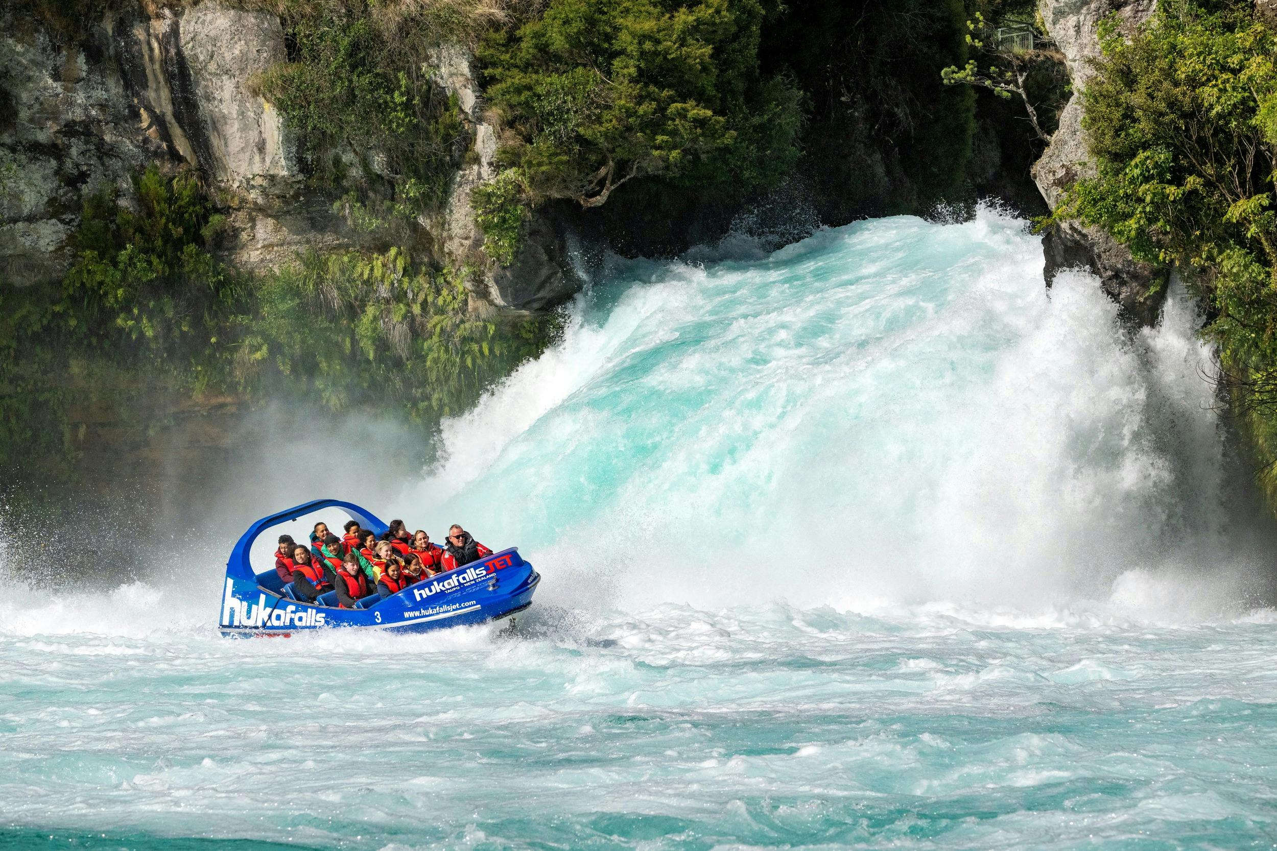 A blue jet boat with passengers approaches a waterfall surrounded by greenery.