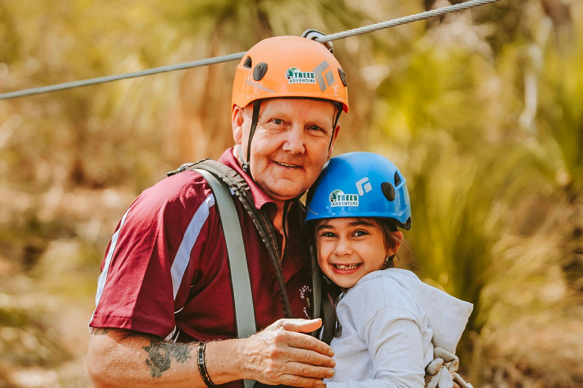 一名成年人和一名儿童戴着头盔和马具，在户外探险公园中微笑着站在一起。
