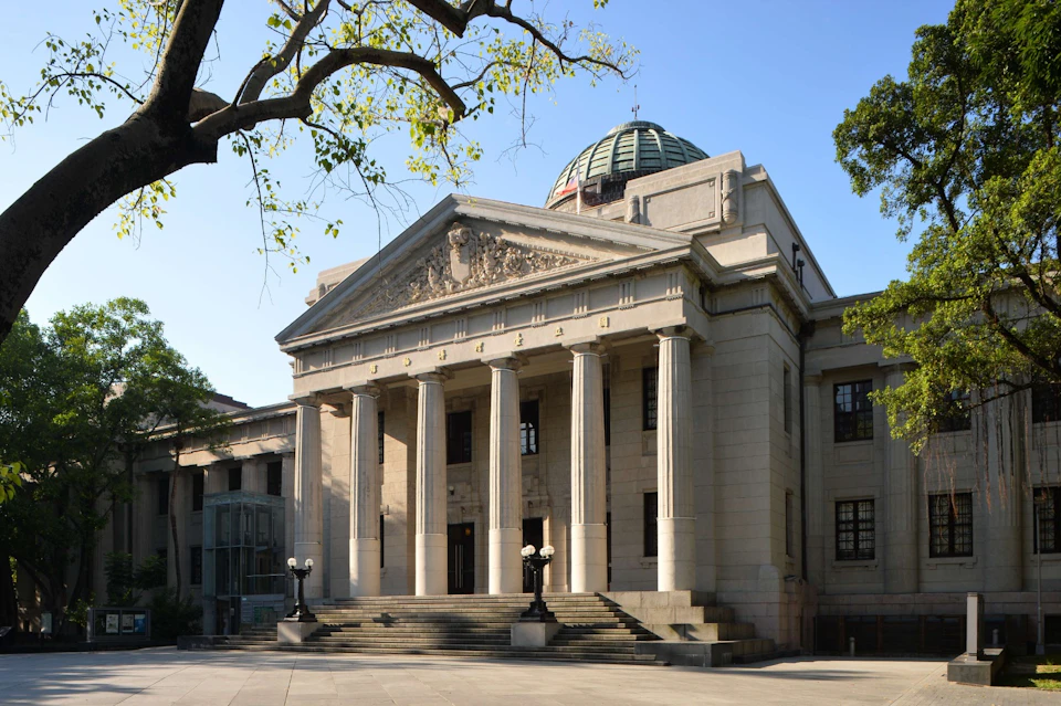 A neoclassical building with tall columns, a pediment, and a domed roof, surrounded by trees under a clear sky.