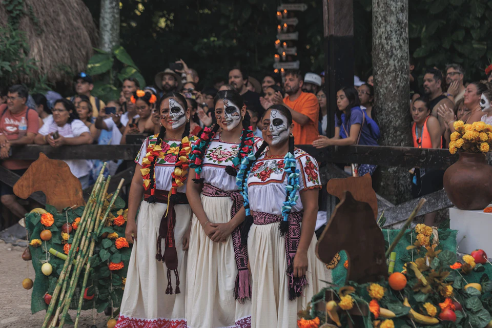 A group of women wearing traditional attire and face paint stand by an altar with marigolds and fruits, surrounded by a crowd.