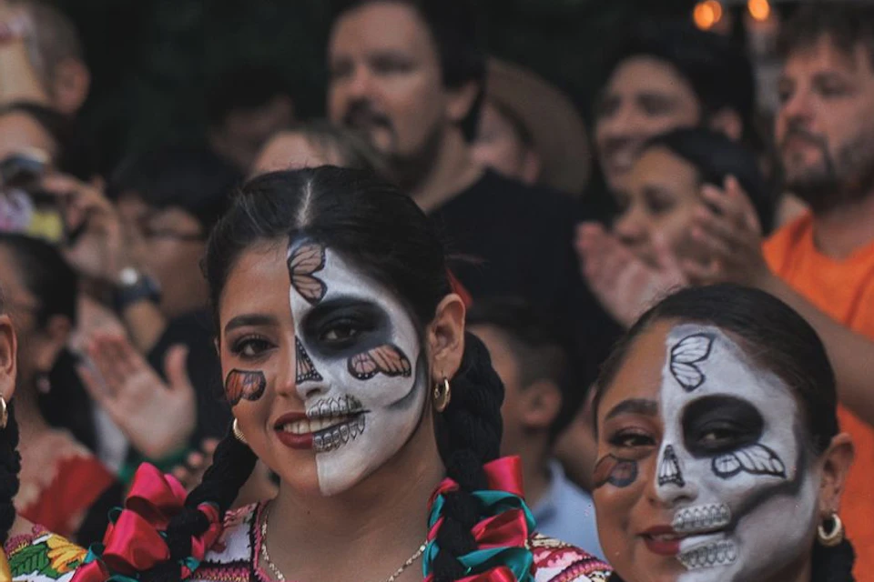 A group of women wearing traditional attire and face paint stand by an altar with marigolds and fruits, surrounded by a crowd.