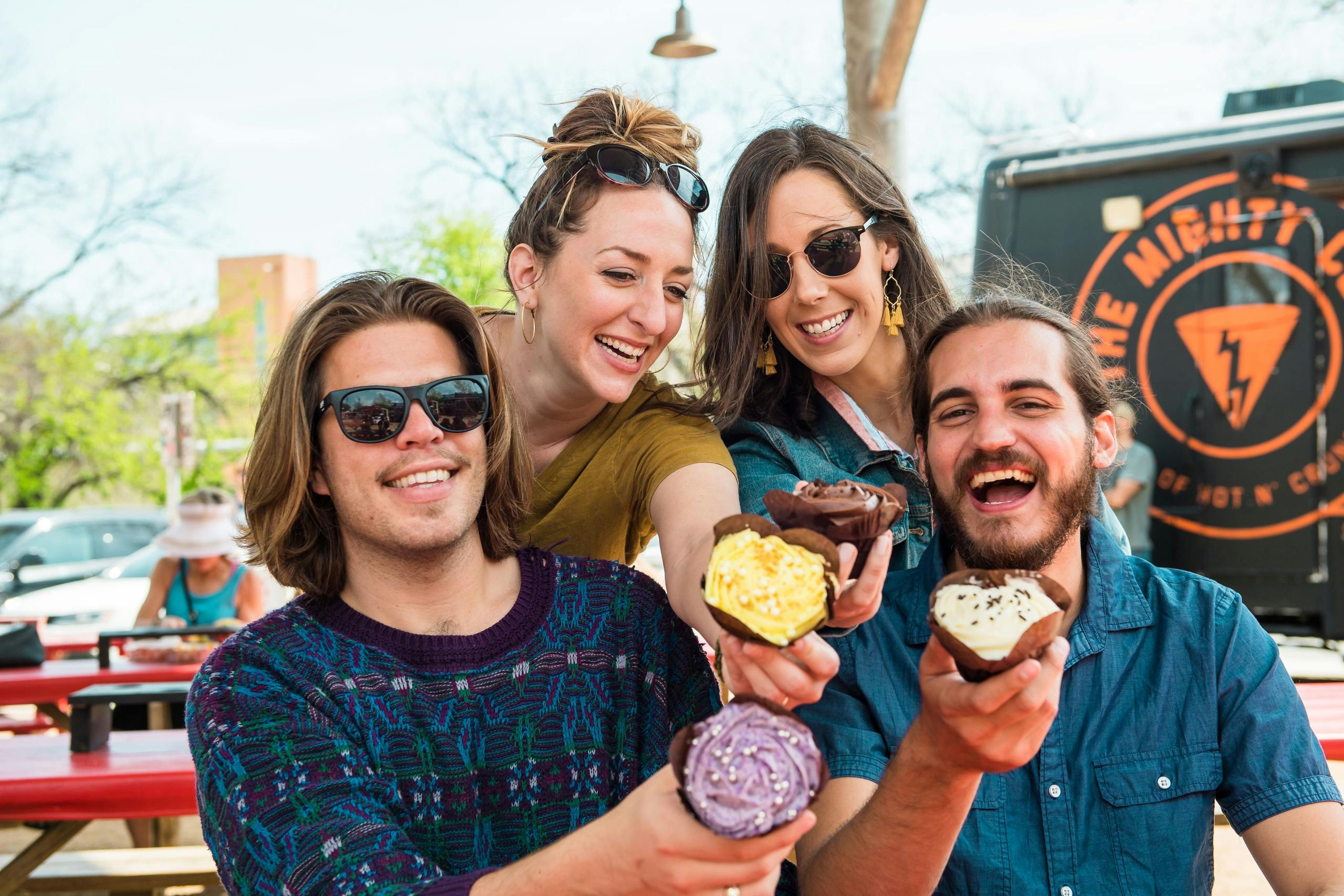 Quatre personnes souriant et tenant des petits gâteaux décorés en plein air par une journée ensoleillée.