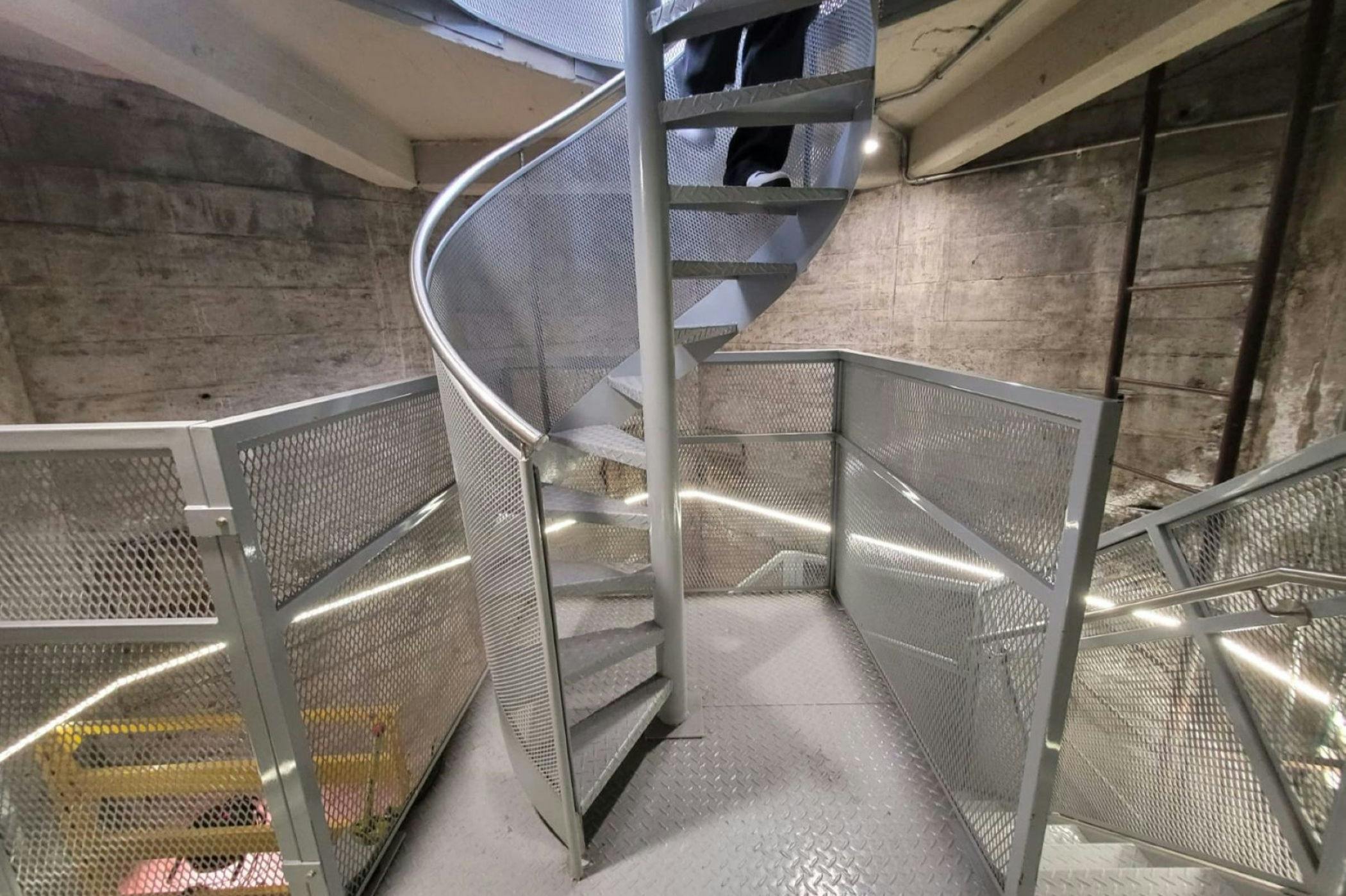 A metallic spiral staircase with mesh railings, set in an industrial interior with concrete walls and visible beams.