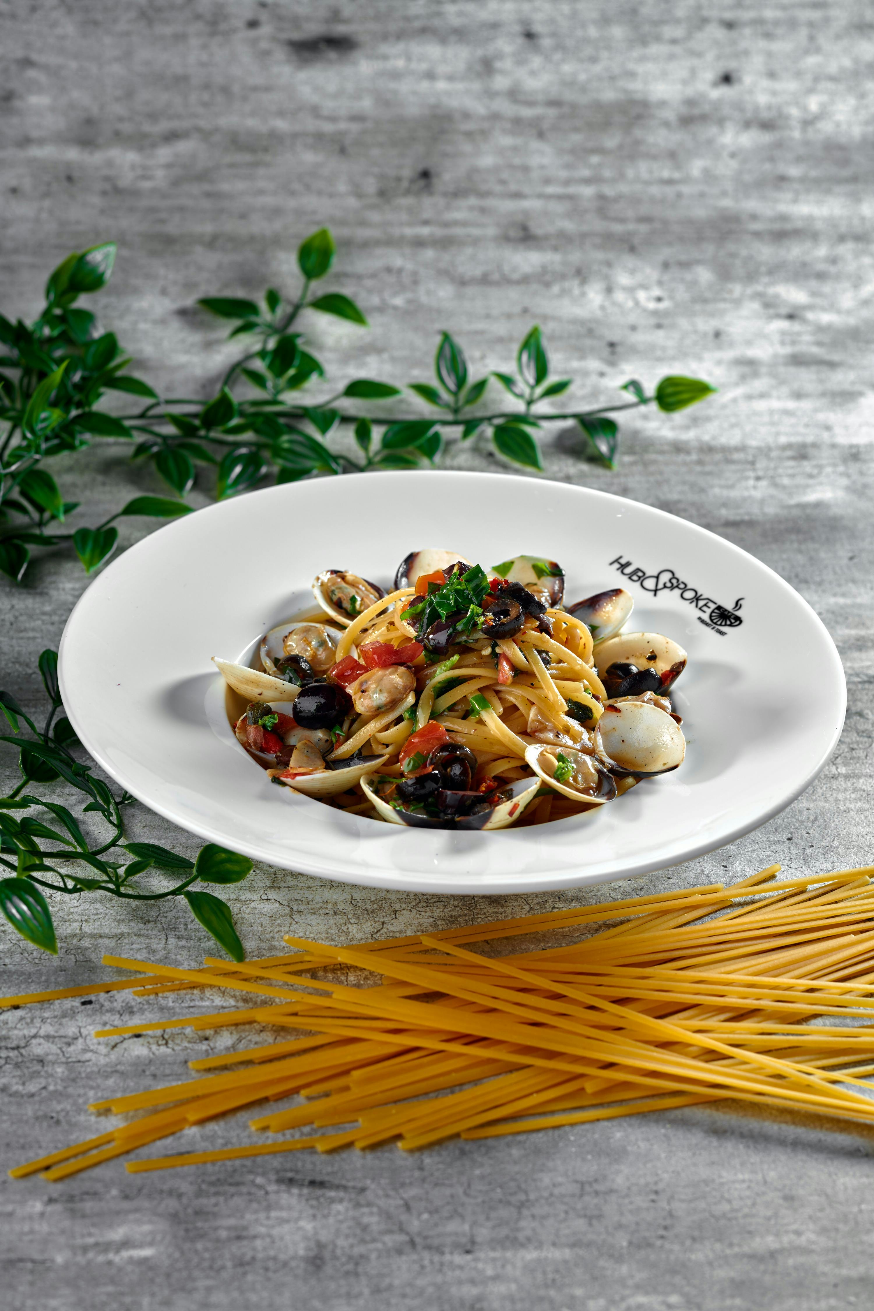 A plate of pasta with clams, olives, and herbs, garnished with parsley, placed on a rustic surface with raw spaghetti nearby.