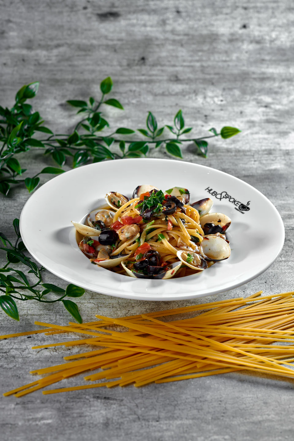 A plate of spaghetti with clams, olives, and herbs, garnished with parsley, beside uncooked spaghetti and green leaves.