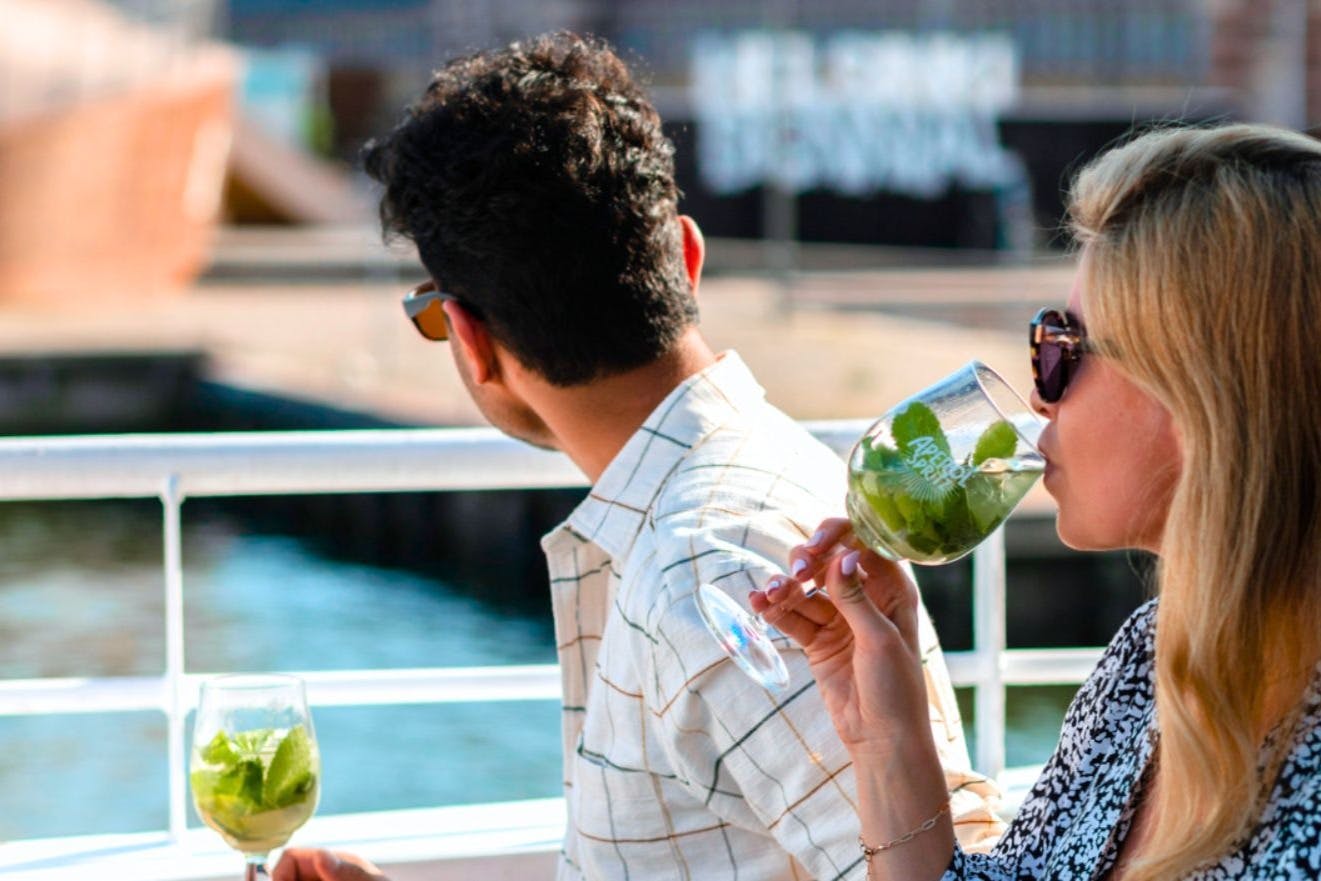 Two people in sunglasses enjoying drinks with green leaves by the water. One faces away while the other sips from a glass.