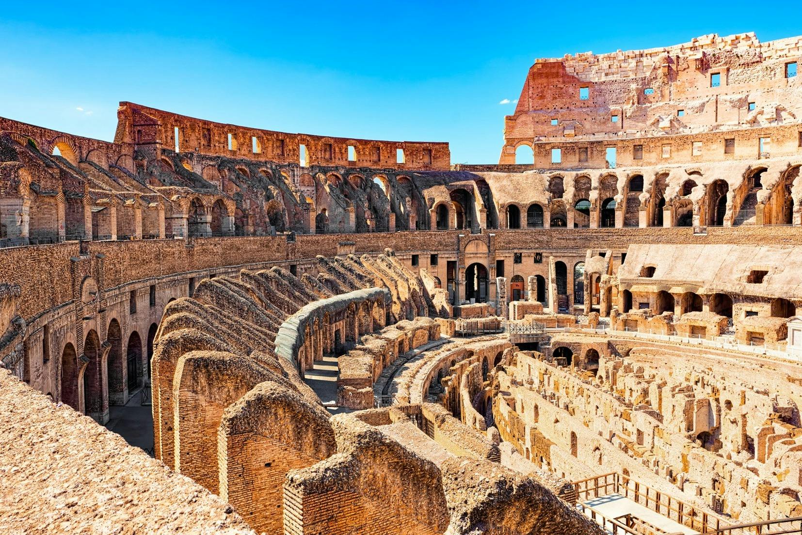 Indvendig udsigt over Colosseum i Rom, der viser dets gamle stenbuer, inddelte siddepladser og delvise ruiner under en blå himmel.