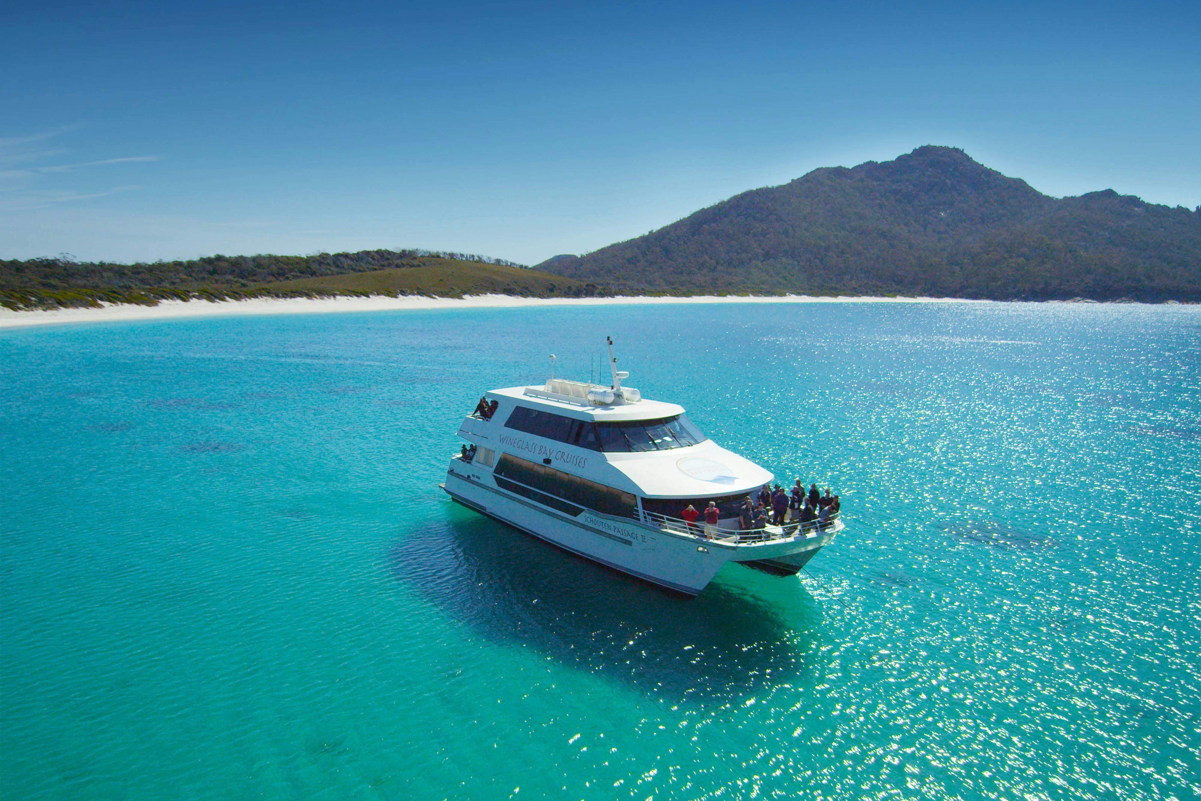A boat with passengers sails on clear turquoise water near a scenic coastline with hills and a sandy beach under a blue sky.
