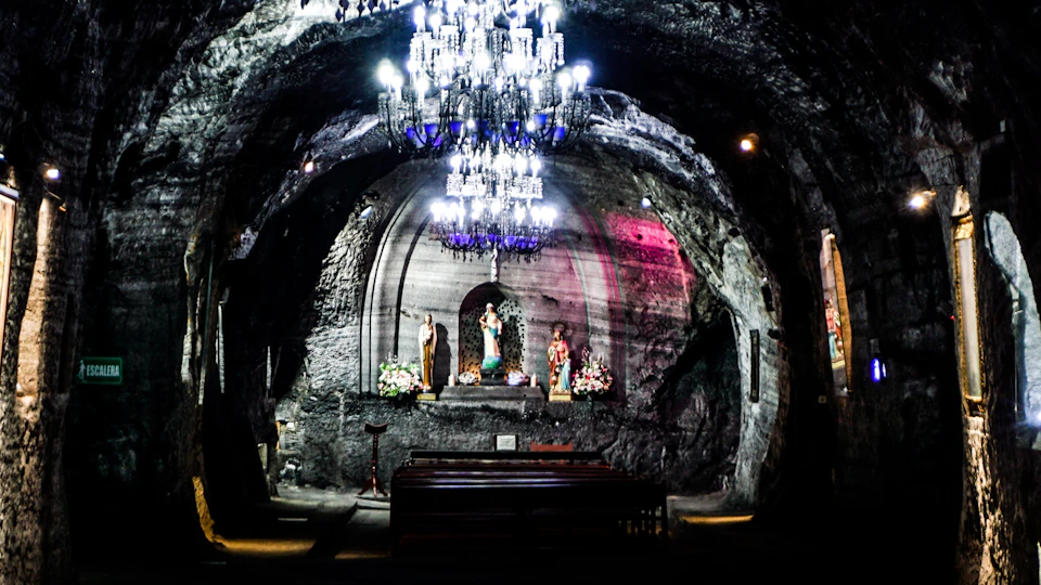 Underground chapel with a lit chandelier, religious statues, floral arrangements, and pews, within a rocky cavern.