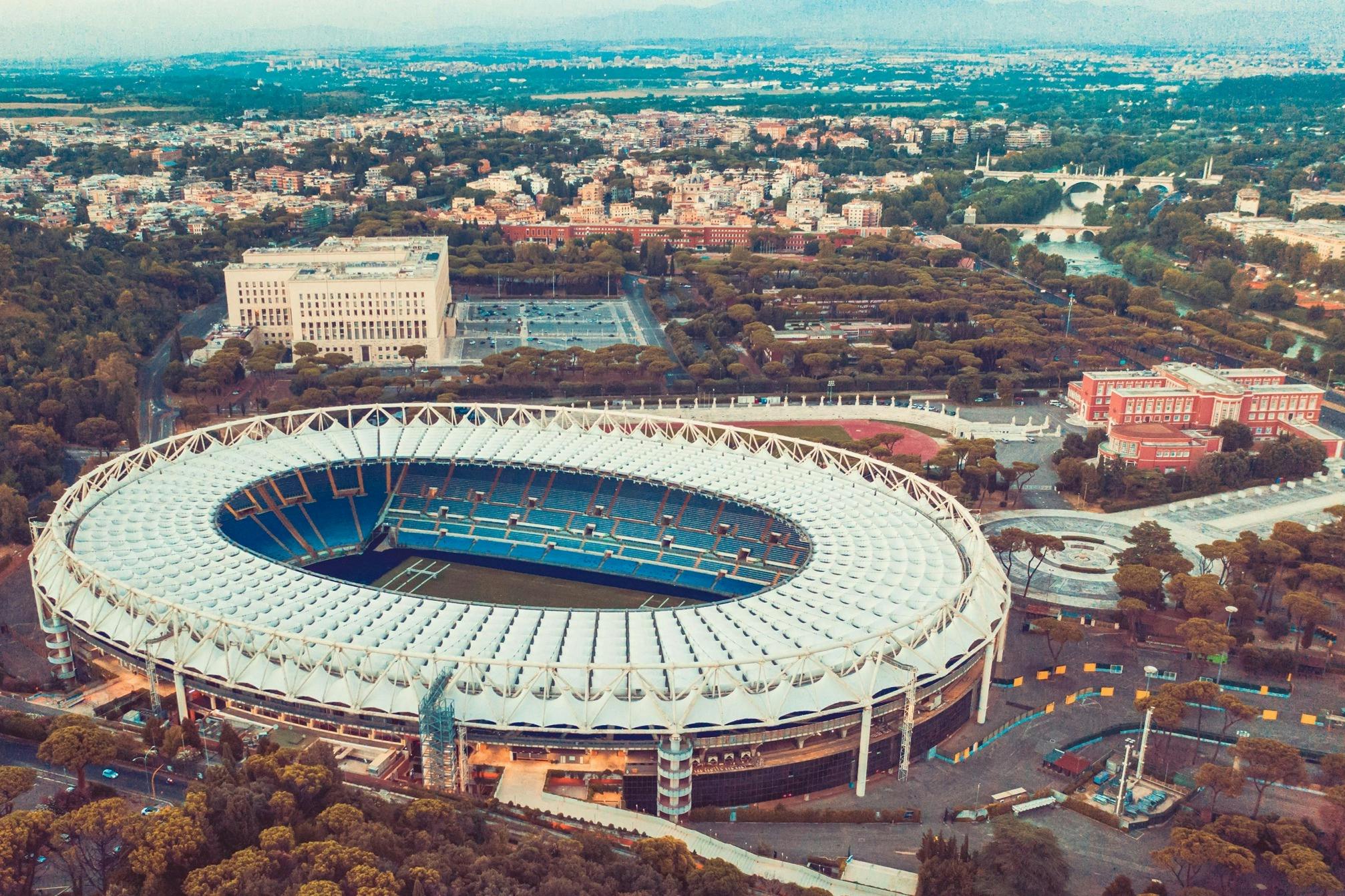 Luchtfoto van een groot stadion met een wit dak omringd door stedelijke gebouwen, groen en bergen in de verte.
